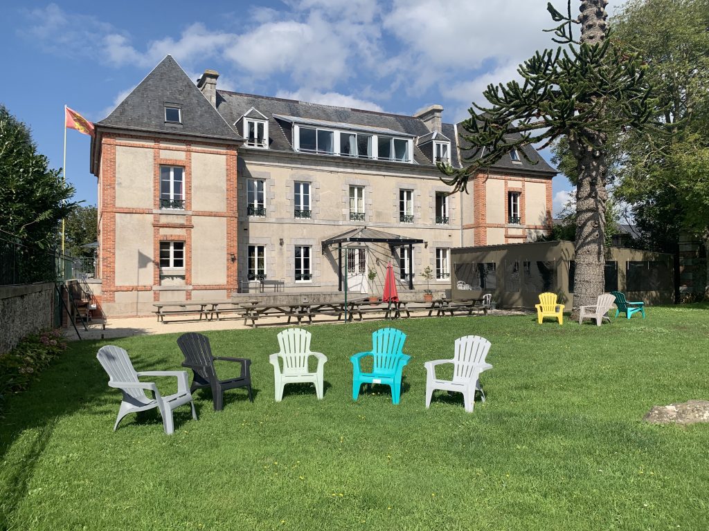 Gîte 50 personnes en Normandie, vu de face avec son jardin et quelques tables et chaises installées dans le parc arboré.